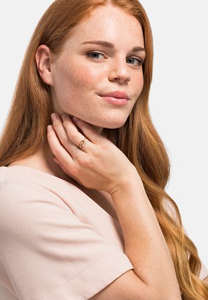 Anello d'oro con design intrecciato indossato sulle dita, caratterizzato da una finitura liscia. Il modello ha lunghi capelli rossi e pelle lentigginosa, su uno sfondo neutro.