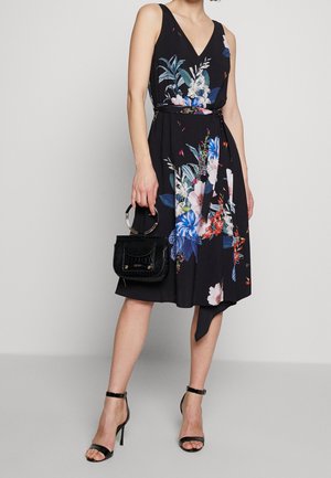 Woman wearing black sleeveless floral dress with asymmetrical hem, holding black handbag with circular handles, and black high-heeled sandals.