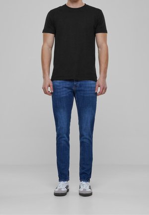 Mann trägt ein schlichtes schwarzes kurzärmeliges T-Shirt, blaue Slim-Fit-Jeans und weiße Sneakers mit schwarzen Streifen, steht vor einem schlichten grauen Hintergrund.
