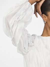 Blusa bianca texturizzata con maniche a sbuffo e volant. Il tessuto è trasparente con un motivo ondulato, presenta un colletto rotondo e bordi morbidi.