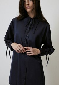 Abito camicia blu navy in tessuto liscio, con un ampio colletto, chiusura a bottoni e dettagli legati sulle maniche. Maniche lunghe e vestibilità ampia.