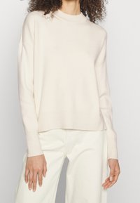 Ljusbeige stickad tröja med hög halsringning, sänkta axlar och kort passform. Mjukt material och ribbade ärmslut framhäver designen.