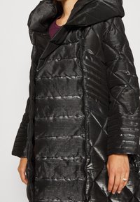 Giacca imbottita nera con finitura lucida, dotata di ampio cappuccio, motivo trapuntato diagonale e chiusura con zip. Maniche testurizzate con dettagli a coste.