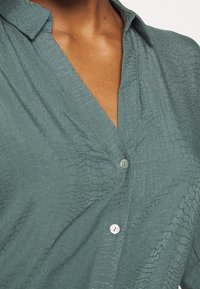 Blouse verte texturée avec un motif reptile, dotée d'un col à revers et d'une fermeture boutonnée. Tissu doux avec un léger éclat.