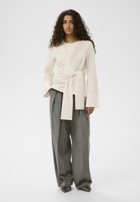 Kreemja wrap-särk pika, laia varrukaga ja esise sidumisega, koos halli triibulise laia jalgadega pükstega ja sõlmega hallide kingadega.