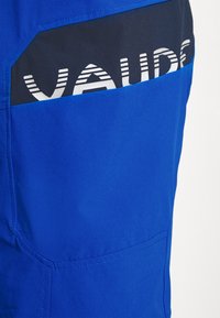 Sinisiä kangashousuja, joissa on tummansinisestä kankaasta valmistettu erottuva tasku, jossa on "VAUDE"-logo valkoisella tekstillä. Housuissa on sileä pinta ja siistit linjat.