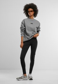 Felpa grigia con un logo rettangolare nero, abbinata a leggings neri e sneakers grigie. La felpa ha maniche larghe e scollo rotondo.