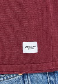 Burgundské pletené tkaniny s bielou nálepkou Jacks & Jones a čiernym textom. Detailný záber s viditeľnou textúrou a švom.