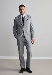 Abito grigio chiaro con design aderente, tessuto strutturato, giacca monopetto e pantaloni coordinati. Completo di camicia bianca e cravatta scura.