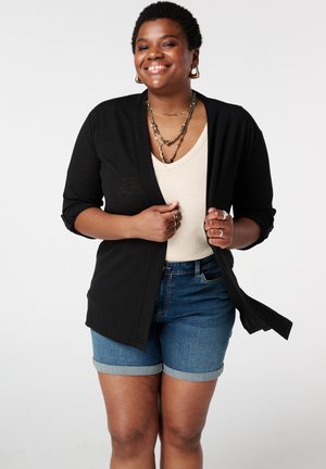 Personne souriante portant des colliers superposés, un cardigan noir sur un haut beige et un short en jean, tenant les bords du cardigan.