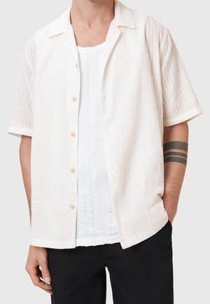 Homme portant une chemise légèrement texturée à manches courtes boutonnée par-dessus un débardeur blanc, un pantalon noir et un tatouage de brassard noir.