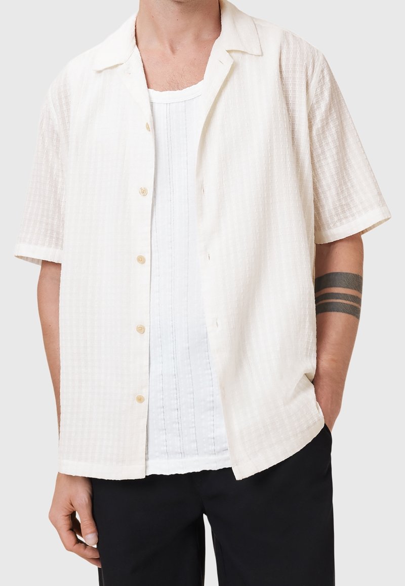 Homme portant une chemise légèrement texturée à manches courtes boutonnée par-dessus un débardeur blanc, un pantalon noir et un tatouage de brassard noir.