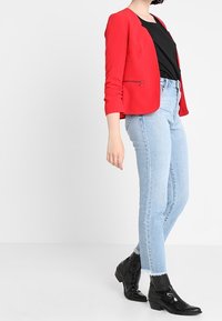 Blazer rouge à manches trois-quarts avec un détail zippé, associé à un haut noir, un jean bleu clair taille haute et des bottines noires.
