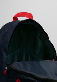 Námorně modrý batoh s červeným zavěšeným madlem, s otevíracím vrškem na zip a texturovanou vnitřní podšívkou ve tmavě zelené barvě.