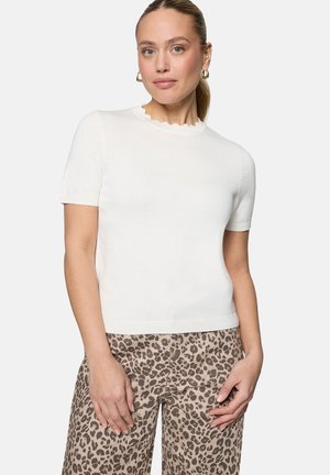 Frau mit goldenen Creolen, die ein weißes kurzärmeliges Oberteil und beige Leopardenmuster-Hosen trägt, vor einem schlichten Hintergrund stehend.