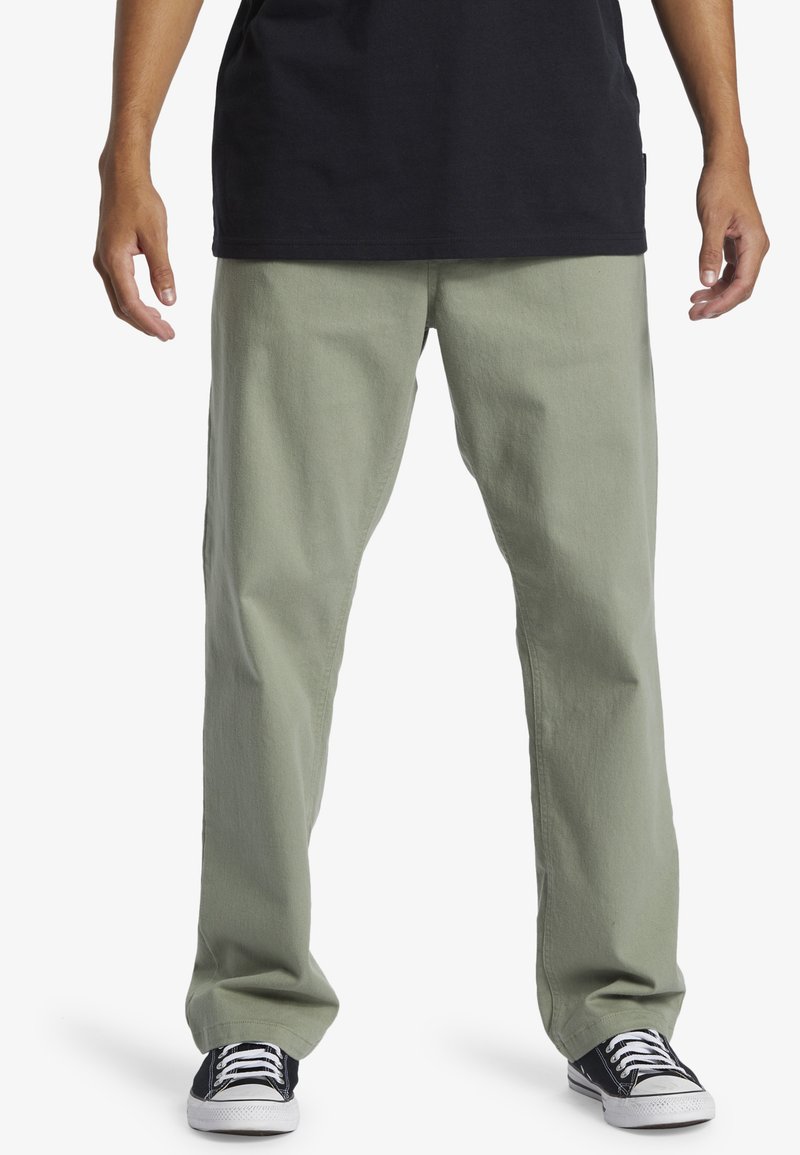 Pantaloni din bumbac de culoare verde măslin deschis, cu o croială relaxată, picioare drepte și buzunare laterale, asortați cu adidași din pânză alb-negru.