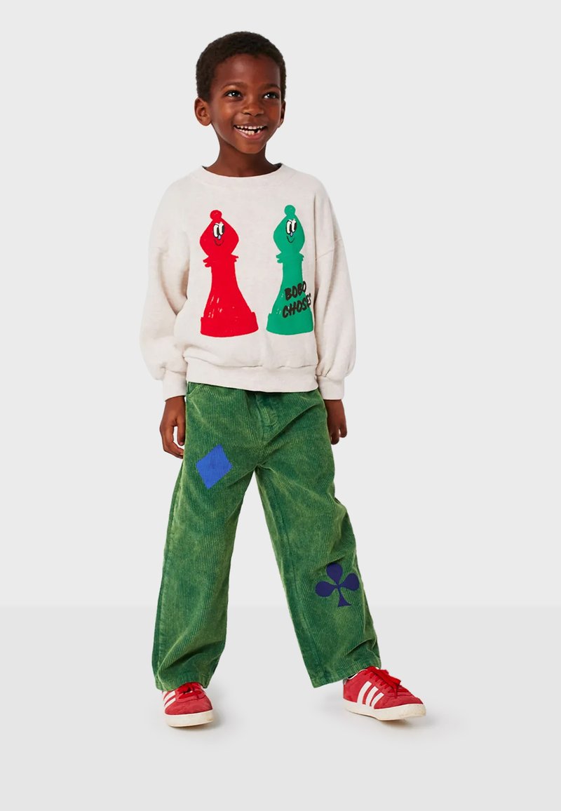Bambino sorridente che indossa una felpa bianca con pedoni a scacchi rossi e verdi, pantaloni verdi con toppe a forma di semi di carte blu e sneaker rosse.