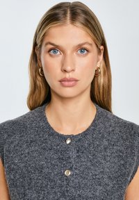 Grau gestrickte Weste mit rundem Halsausschnitt, kurzen Ärmeln und zwei silbernen Knopfakzenten an der Vorderseite. Die weiche Textur wirkt gemütlich.