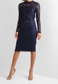 Marinblå spetsklänning med långa ärmar, blommigt mönster, figurnära silhuett, längd nedanför knäna, matchad med strap-line metalliska klackar.