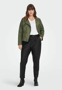 Chaqueta de motocicleta de ante sintético verde, camiseta blanca con escote en V, pantalones de cuero sintético negros y botines. Presenta detalles con cremallera y corte relajado.