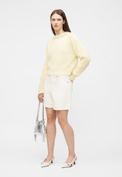 Γυναίκα που φοράει απαλό κίτρινο χνουδωτό πουλόβερ και λευκό σορτς, κρατώντας ασημί τσάντα, στέκεται με το ένα χέρι στην τσέπη και ουδέτερη έκφραση.