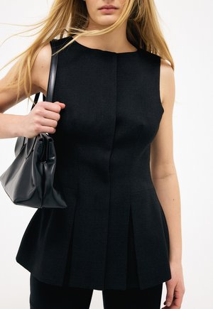 Kobieta w czarnym plisowanym topie bez rękawów, trzymająca czarną skórzaną torebkę na ramieniu, z długimi blond włosami.