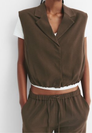 Femme portant un gilet court à rayures marron avec revers, sur un t-shirt blanc, et un pantalon marron assorti à cordon de serrage, les mains dans les poches.