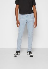 Jeans rectos de color azul claro con dos bolsillos frontales, un bolsillo trasero y un botón marrón. Llevados con una camiseta negra y zapatillas en blanco y negro.