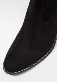 Botín de ante negro hasta el tobillo con punta afilada, que presenta un acento gris cosido a lo largo de la suela y una textura suave.