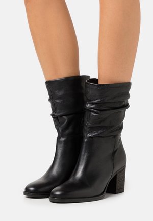 Czarne skórzane kozaki do połowy łydki z luźną cholewką, zaokrąglonym noskiem i zgrupowanymi słupkowymi obcasami, noszone na gołych nogach na jednolitym tle.