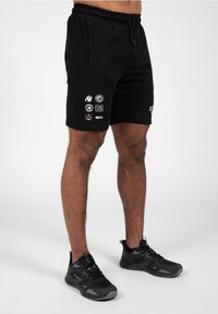 Zwarte shorts met elastische tailleband, voorzien van witte grafische prints, waaronder iconen en cijfers. Gedragen met zwarte sportschoenen.