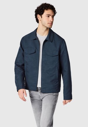 Junger Mann trägt eine marineblaue Jacke über einem hellgrauen Hemd und hellgrauen Jeans, geht und schaut seitlich vor einem einfarbigen Hintergrund.