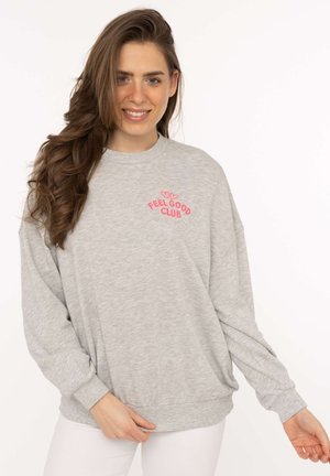 Lächelnde Frau mit langen braunen Haaren trägt ein hellgraues Sweatshirt mit rosa "FEEL GOOD CLUB"-Text und Herzmotiv auf der Brust.
