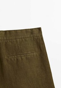 Gros plan sur la ceinture d’un pantalon en lin vert olive avec une poche passepoil horizontale unique sur fond blanc.