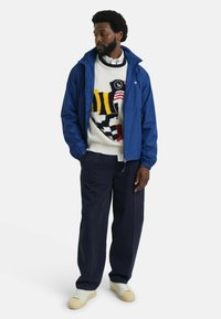 Chaqueta de nylon azul sobre un suéter con un patrón, combinada con pantalones azul marino y zapatillas. La chaqueta tiene una capucha ajustable y puños elásticos.
