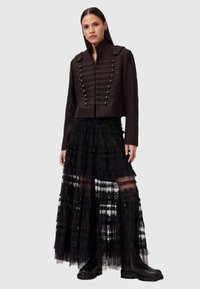 Chaqueta recortada de mezcla de lana negra con adornos de botones al estilo militar, combinada con una falda larga de encaje negro en capas con volantes. Botas negras.