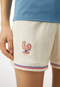 Baltas sporta šorti ar sietiņveida tekstūru, ar gaiļa simbolu un zem tā "FFF". Zili un sarkani svītriņas apakšmalā piešķir detaļu.