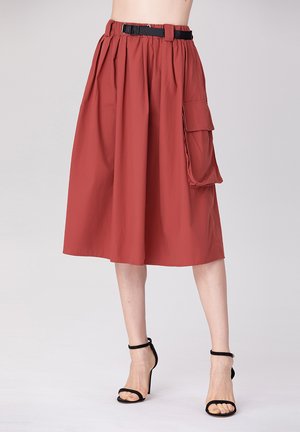Jupe midi rouge en tissu léger avec des plis, poche latérale et taille noire. Associée à des talons noirs à lanières.