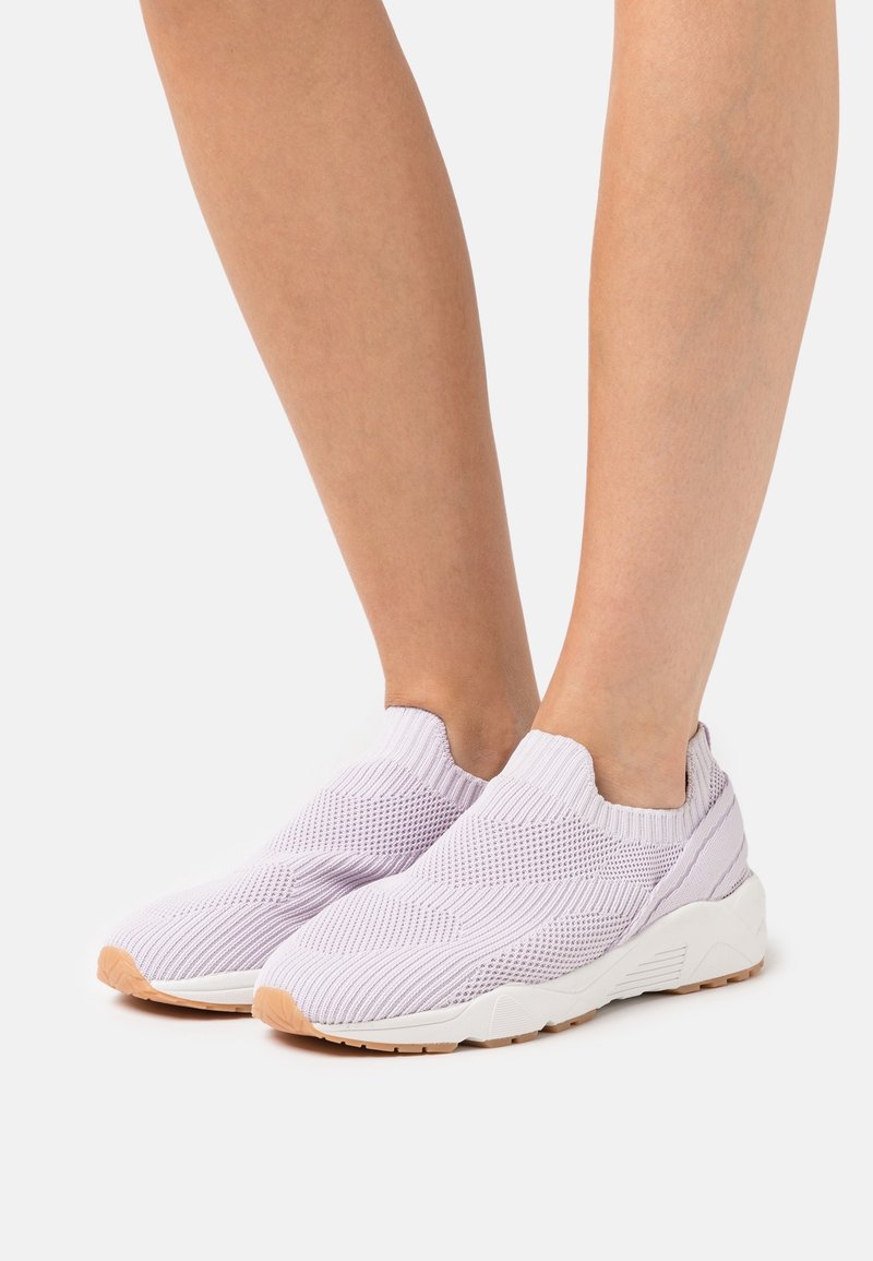 Pieds portant des baskets slip-on tricotées violet clair avec des semelles blanches et des embouts en gomme sur un fond uni.