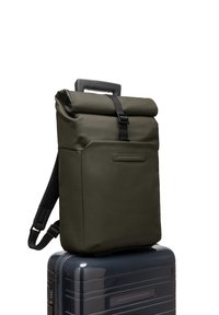 Sac à dos à rabat en vert olive, fabriqué en matériau durable, avec une poche avant, une sangle noire et une poignée robuste sur le dessus. Positionné sur une valise.