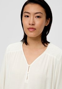 Tunique blanche avec un décolleté en V, des détails de boutons et un smocking texturé aux épaules. Fabriquée en tissu léger.
