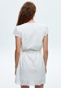 Vit kort klänning med en blommig ögla-design, vågformad nederkant, korta ärmar och ett tygbälte i midjan.