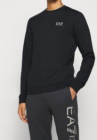 Felpa nera a maniche lunghe in cotone, con scollo rotondo e piccolo logo bianco sul petto a sinistra. Tessuto liscio.