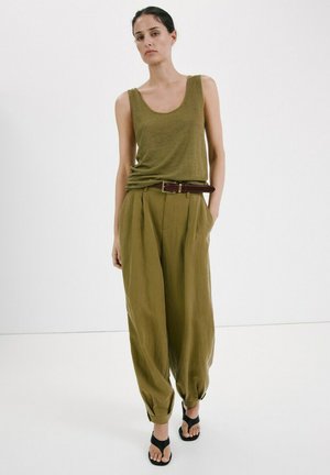 Žena v olivovo zelenej tielkovej blúzke bez rukávov a vo výrazných širokých nohaviciach s vysokým pásom a opaskom, stojaca s jednou rukou v kapsách na bielom pozadí.