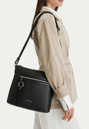 Mujer con chaqueta beige lleva un bolso de hombro de cuero negro con bolsillo delantero con cremallera y detalle de anillo plateado.