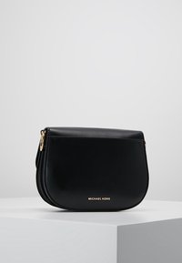Musta nahkainen Michael Kors olkapussukka, jossa on kaarevat reunat ja kultasävytteinen varustus, esillä valkoisella alustalla vaaleanharmaata taustaa vasten.