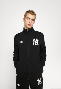 Sort lynlås sweatshirt lavet af blødt stof, med høj krave, frontlommer og et fremtrædende hvidt New York Yankees-logo.