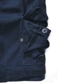 Pantalonii de culoare bleumarin au un buzunar lateral cu un nasture, o cusătură vizibilă și un eticheta mică neagră cu un „B” alb. Materialul texturat pare rezistent.