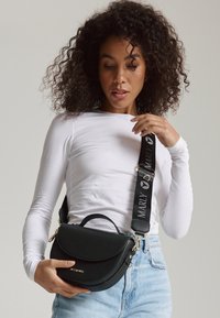 Sac à bandoulière en cuir noir avec une surface texturée, une forme arrondie, des accents dorés et une sangle réglable avec logo.