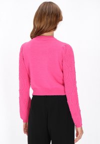 Fuchsia sweater met geribbelde halslijn, textuur kabelbrei mouwen versierd met kleine pailletjes en een cropped fit. Zwarte broek zichtbaar eronder.